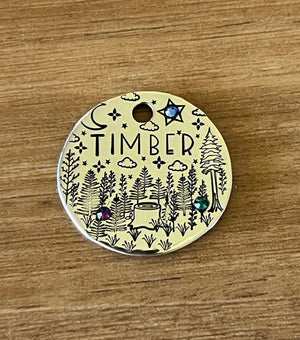 A round metal pet tag with 'TIMBER' written on it, featuring an illustration of a forest, trees, a crescent moon, and stars, adorned with crystal jewels.