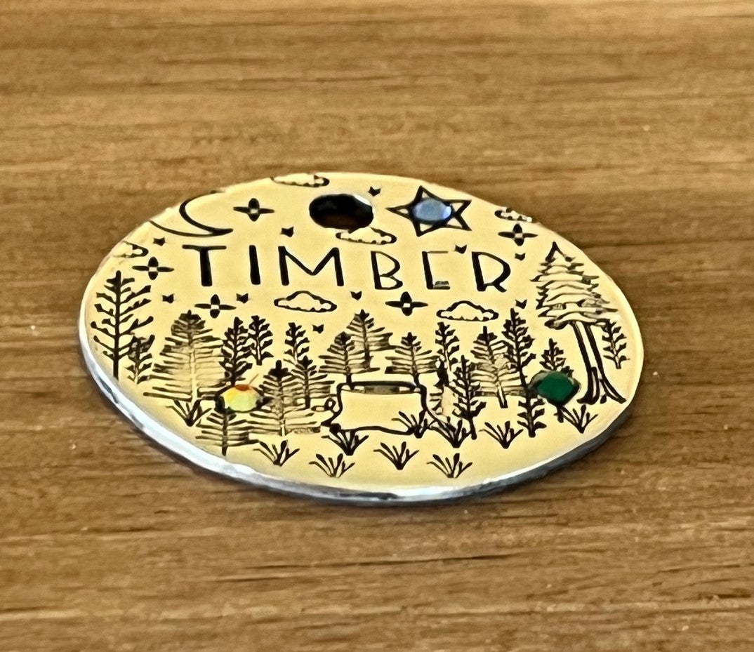 A round metal pet tag with 'TIMBER' written on it, featuring an illustration of a forest, trees, a crescent moon, and stars, adorned with crystal jewels.