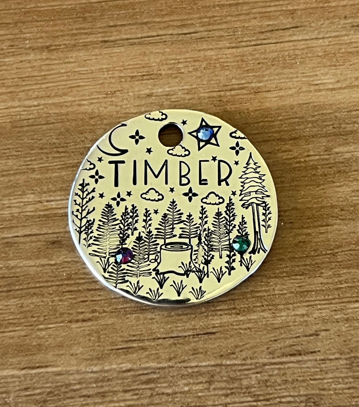 A round metal pet tag with 'TIMBER' written on it, featuring an illustration of a forest, trees, a crescent moon, and stars, adorned with crystal jewels.