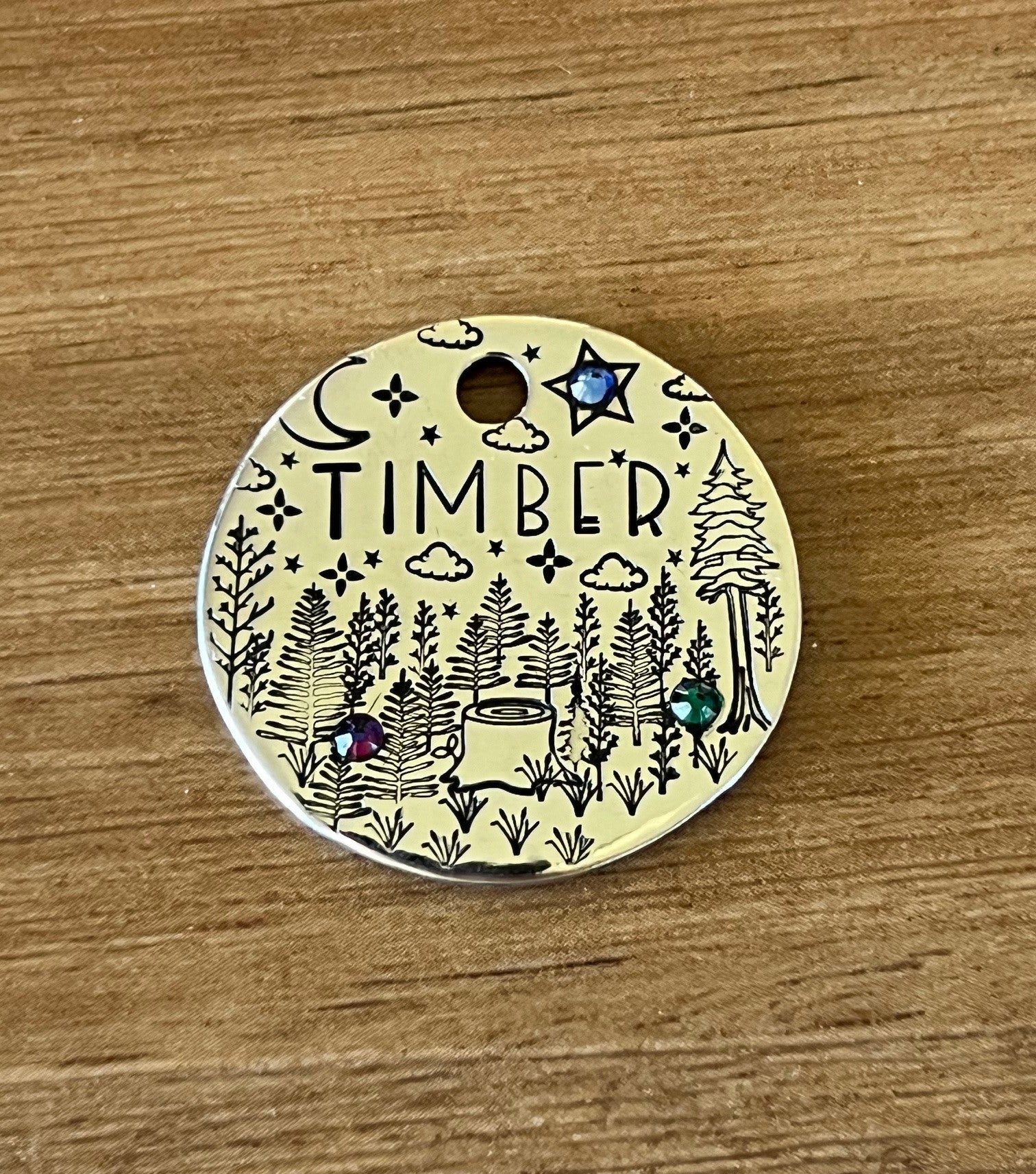 A round metal pet tag with 'TIMBER' written on it, featuring an illustration of a forest, trees, a crescent moon, and stars, adorned with crystal jewels.